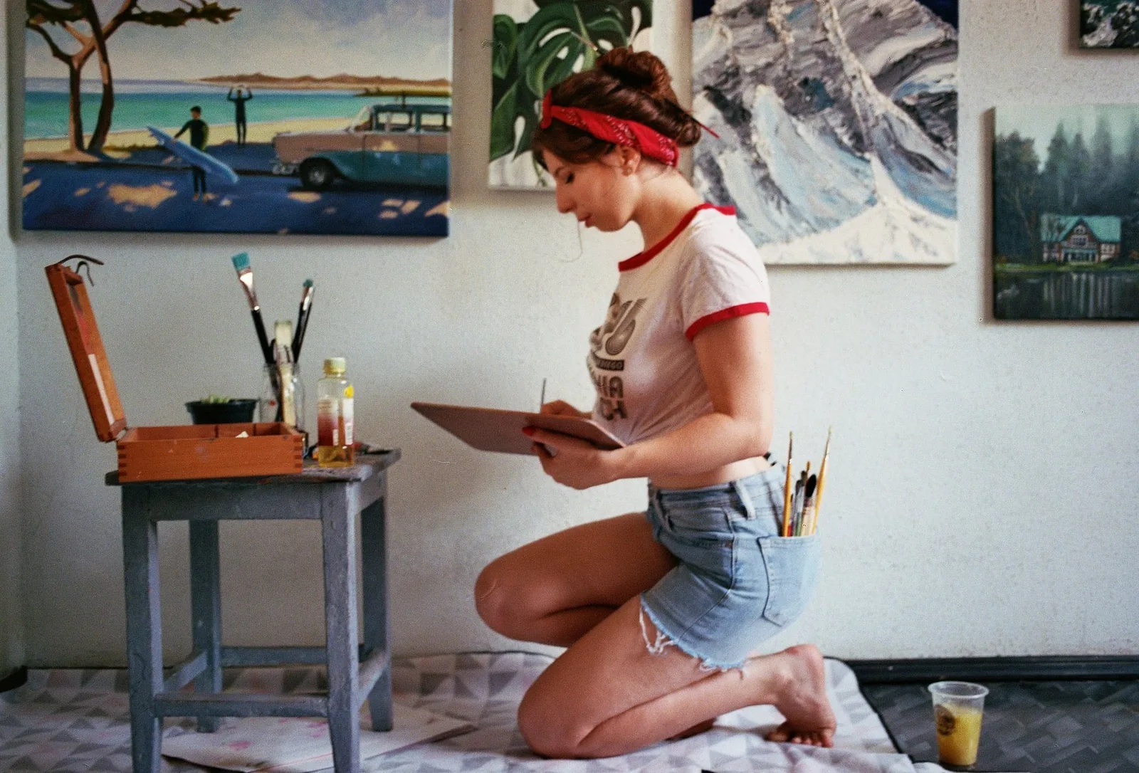 Kate kneeling on the studio floor with a palette, her paintings on the wall behind her